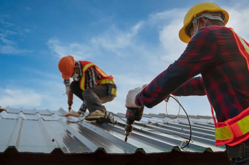 Two roofers in hard hats and hi-vis vests installing a metal roof under a blue sky