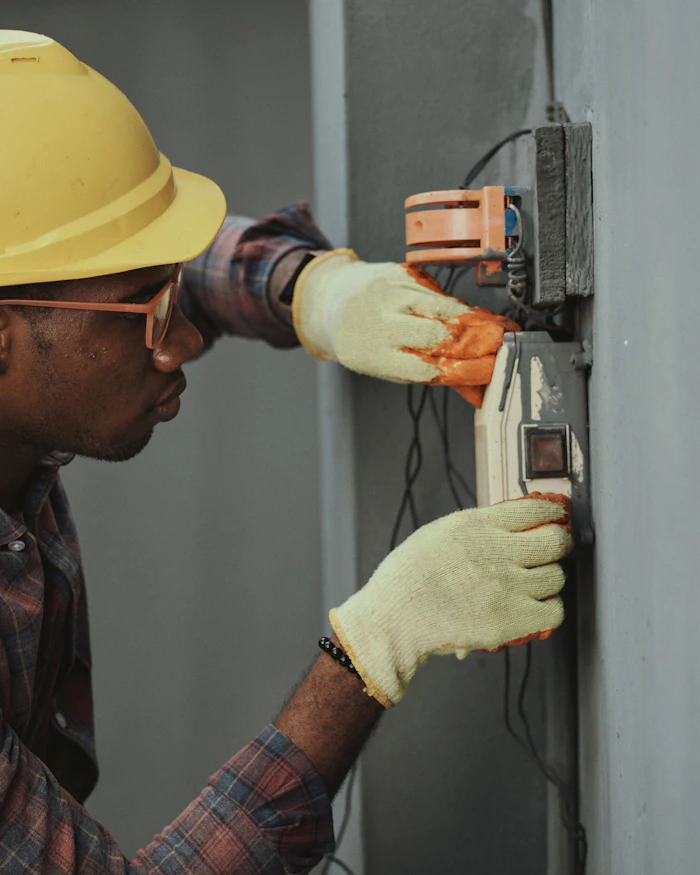 An electrician in a hard hat installing wiring in a wall-mounted box.